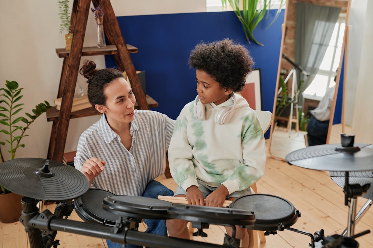 A woman guides a boy with curly hair in learning electronic drums in a cozy indoor setting.