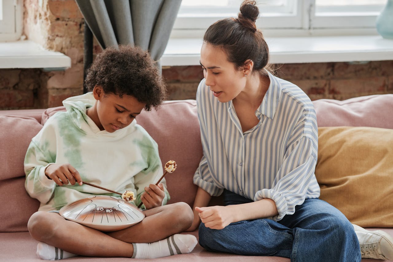 A boy playing a steel tongue drum with guidance from a female instructor on a sofa.